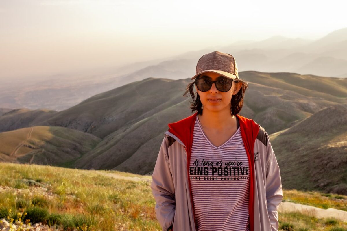 A woman wearing sunglasses and a cap stands in front of a scenic mountain landscape during sunset.